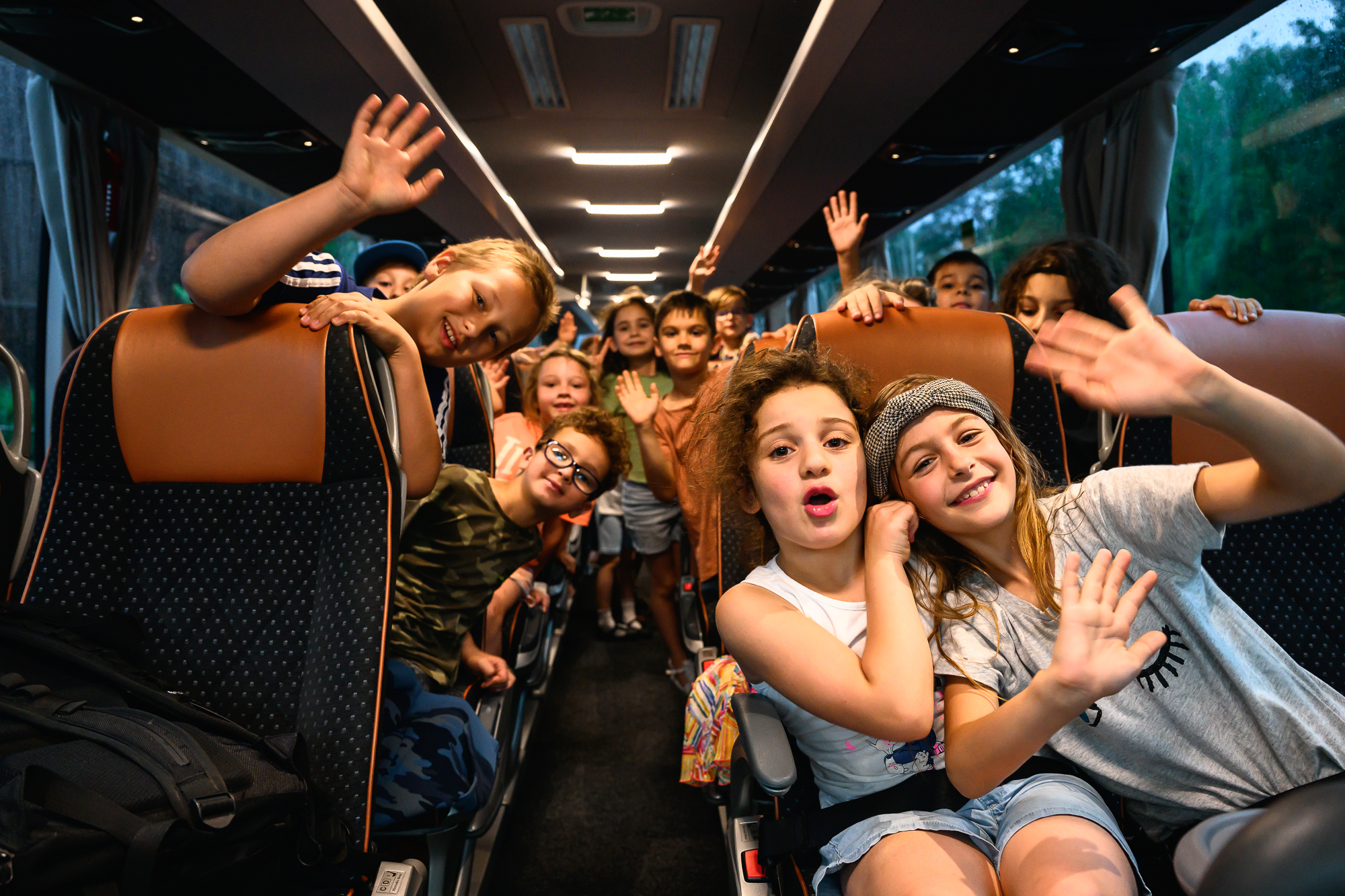 Blick in den Gang durch die Sitzreihen in einem Bus. Die Kinder beugen sich in die Gangmitte und winken in die Kamera. Sie sind so 5-10 Jahre alt.